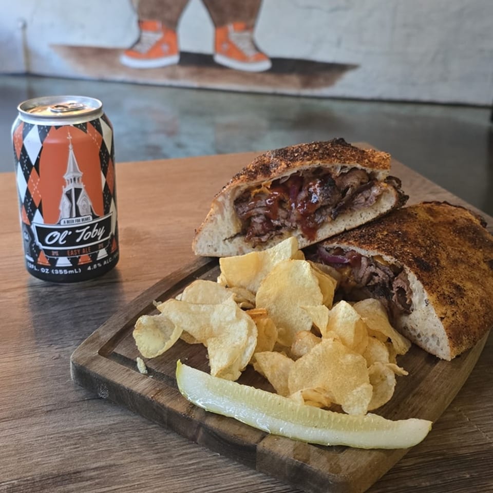 A calzone with chips and a pickle spear on a wooden board, next to an "Ol' Toby" beer can.